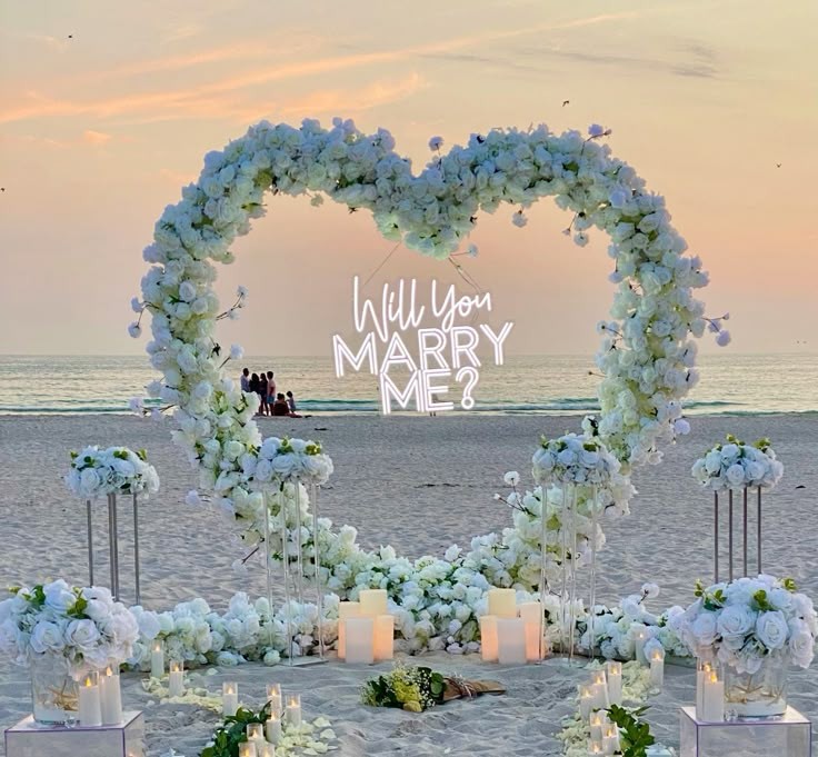 Beach proposal scene with white floral heart arch and candles